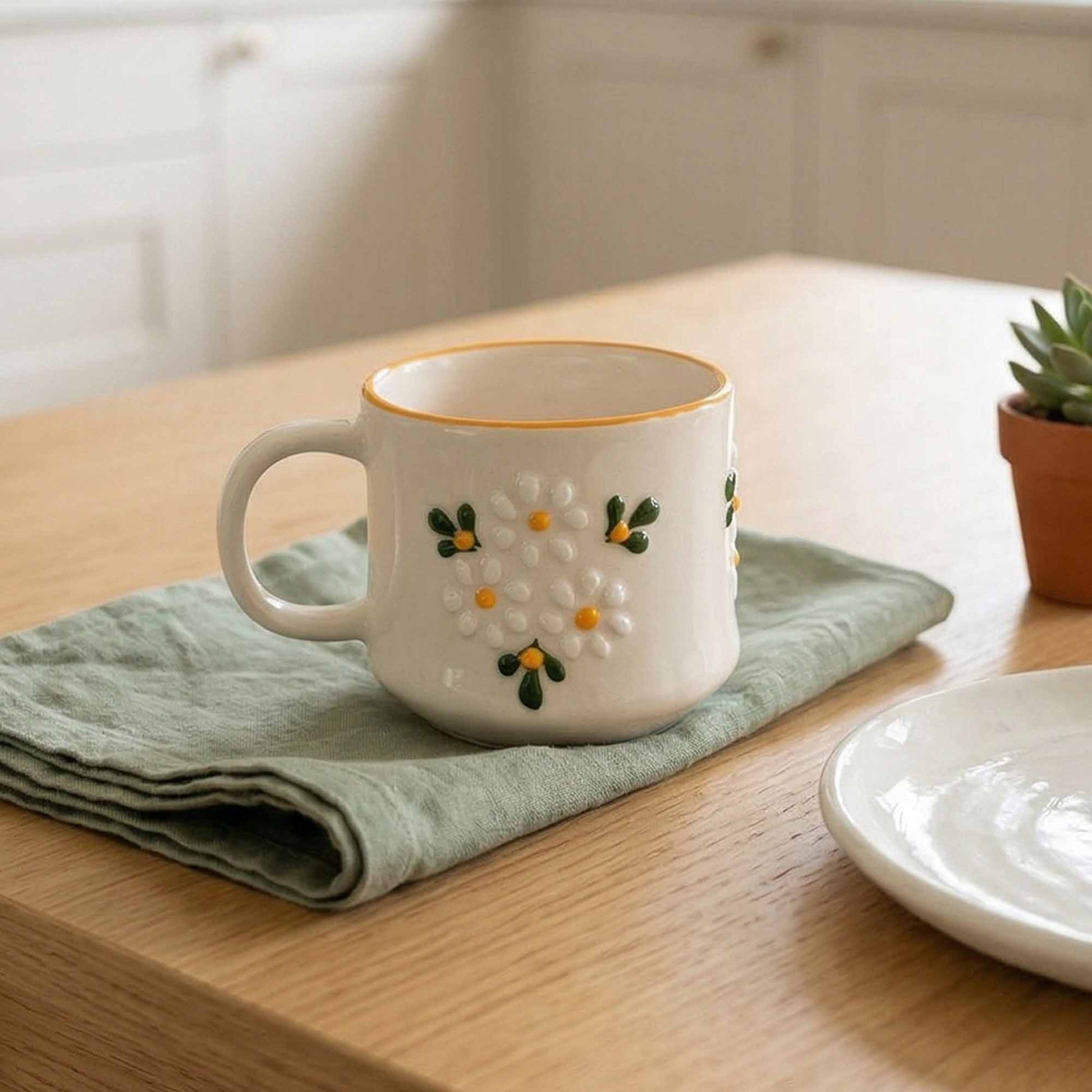 Decorative ceramic mug with floral design on a wooden table with a green cloth and small plant.