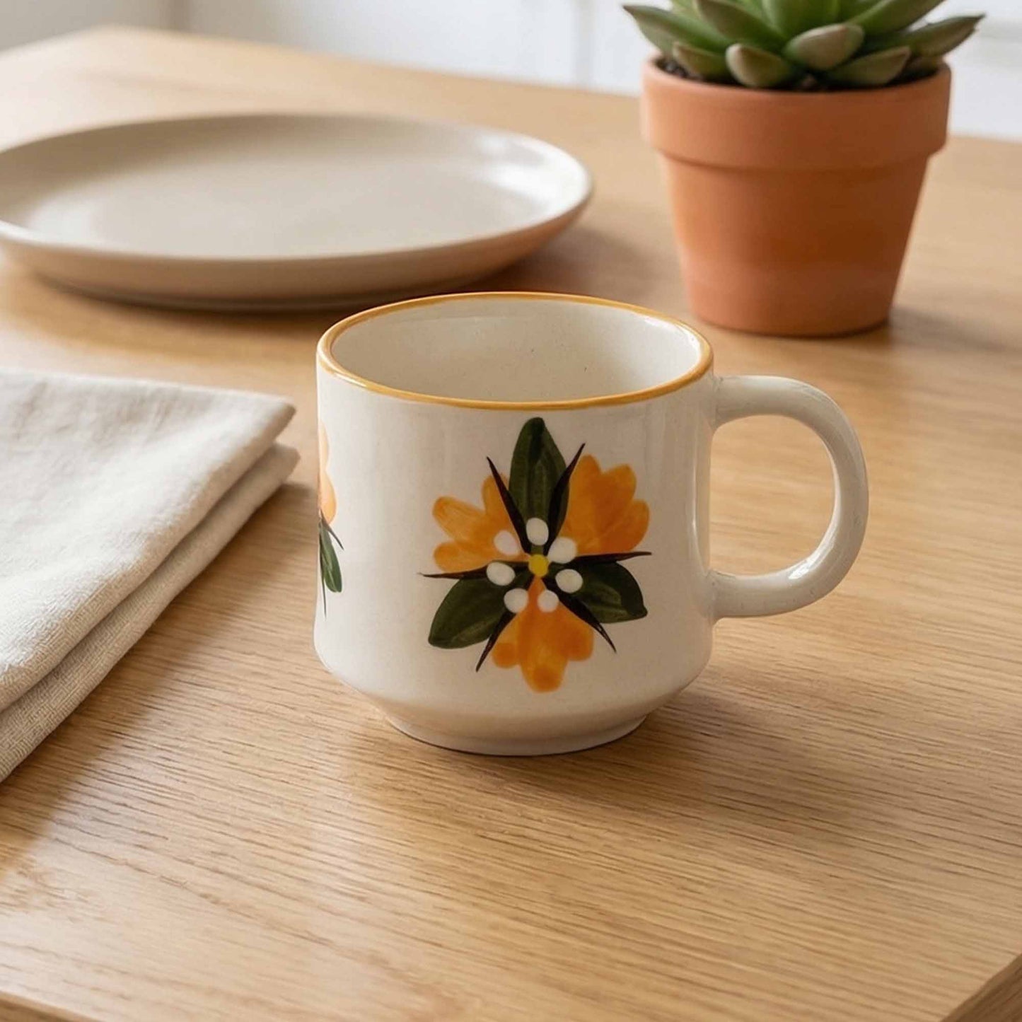Mug with floral design on a wooden table with a plate, napkin, and potted plant in the background.