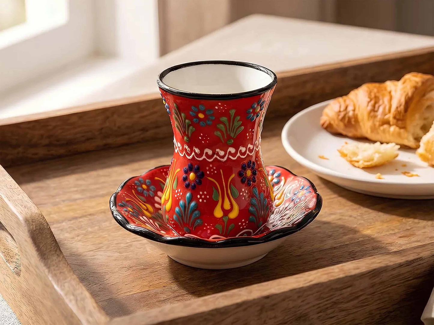 Decorative red cup with floral patterns on a wooden tray with a croissant.