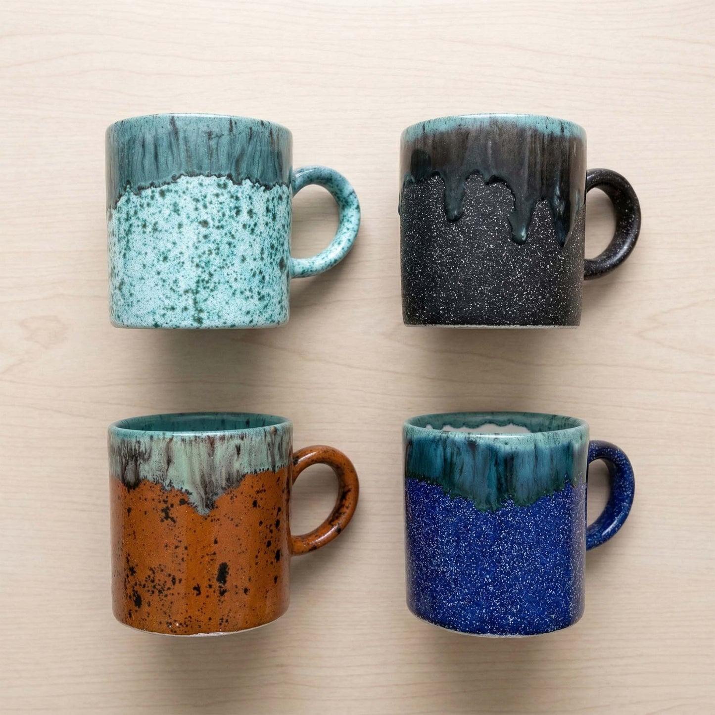 Four ceramic mugs with speckled glaze in different colors on a beige surface.