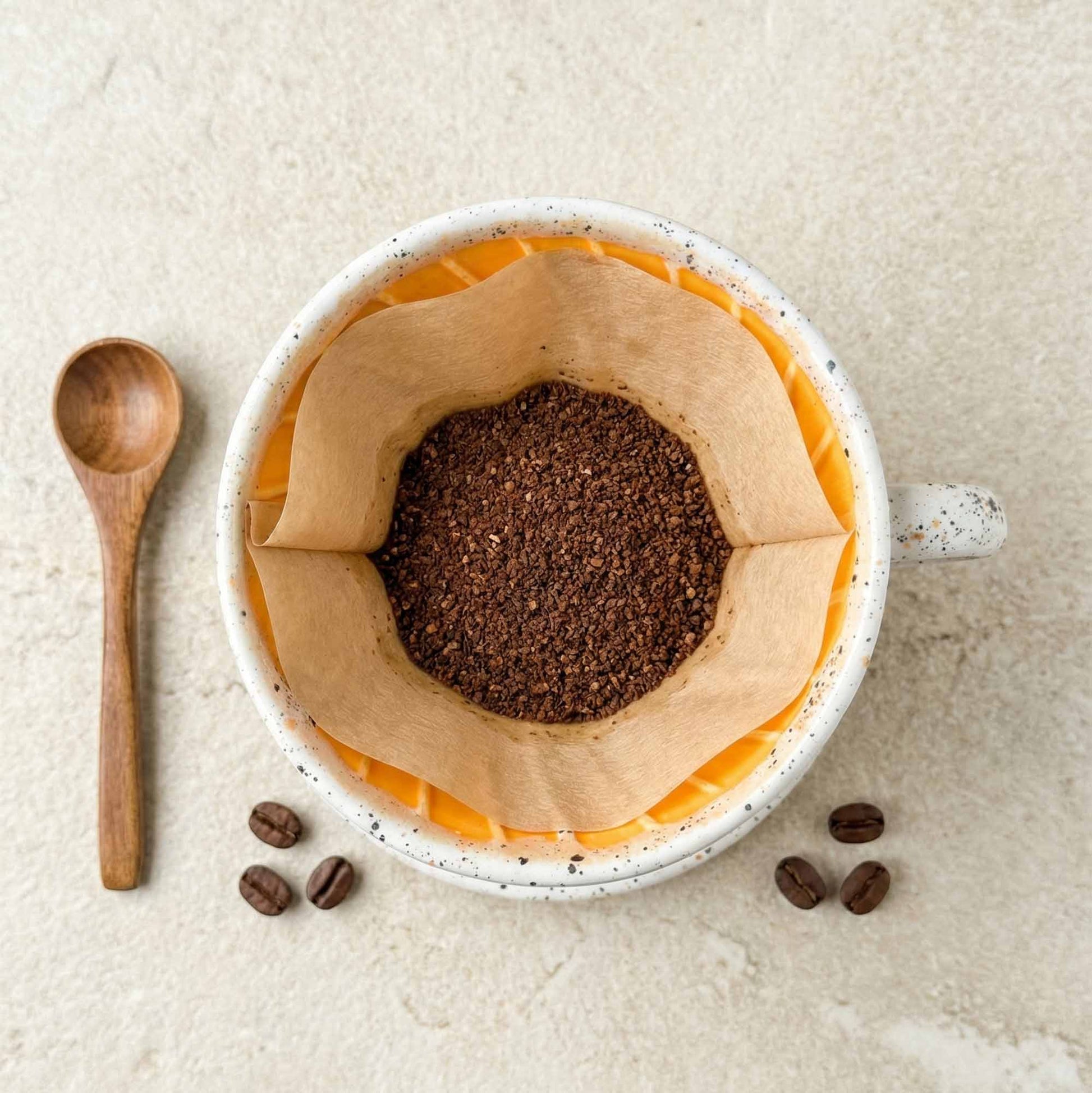 Coffee grounds in a paper filter inside a mug with wooden spoon and coffee beans on a light background