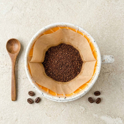 Coffee grounds in a paper filter inside a mug with wooden spoon and coffee beans on a light background