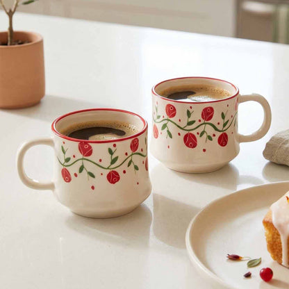 Two ceramic mugs with floral designs filled with coffee on a light surface, accompanied by a plate of pastries.