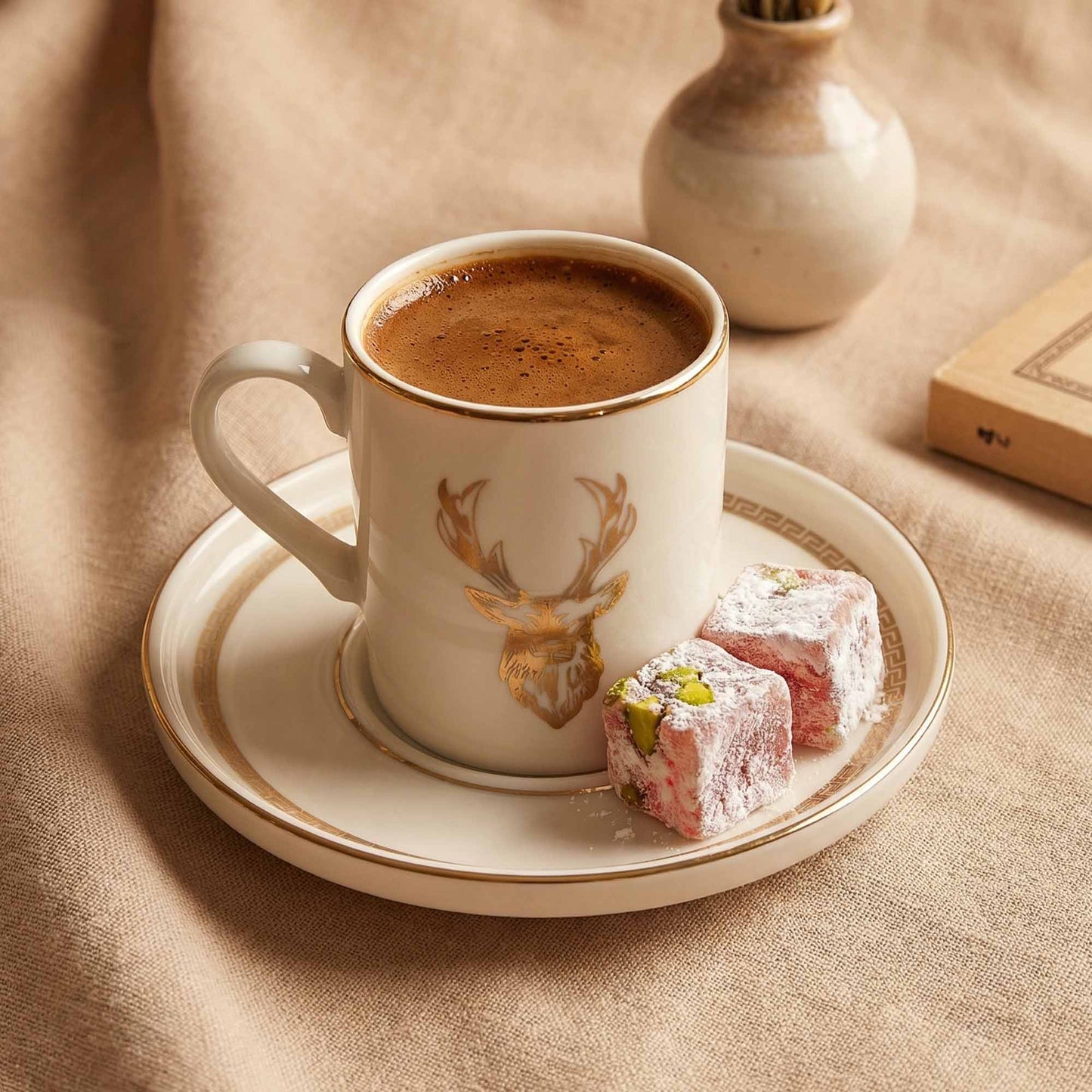 Cup of coffee with a deer design on a saucer, accompanied by Turkish delight on a beige fabric background.
