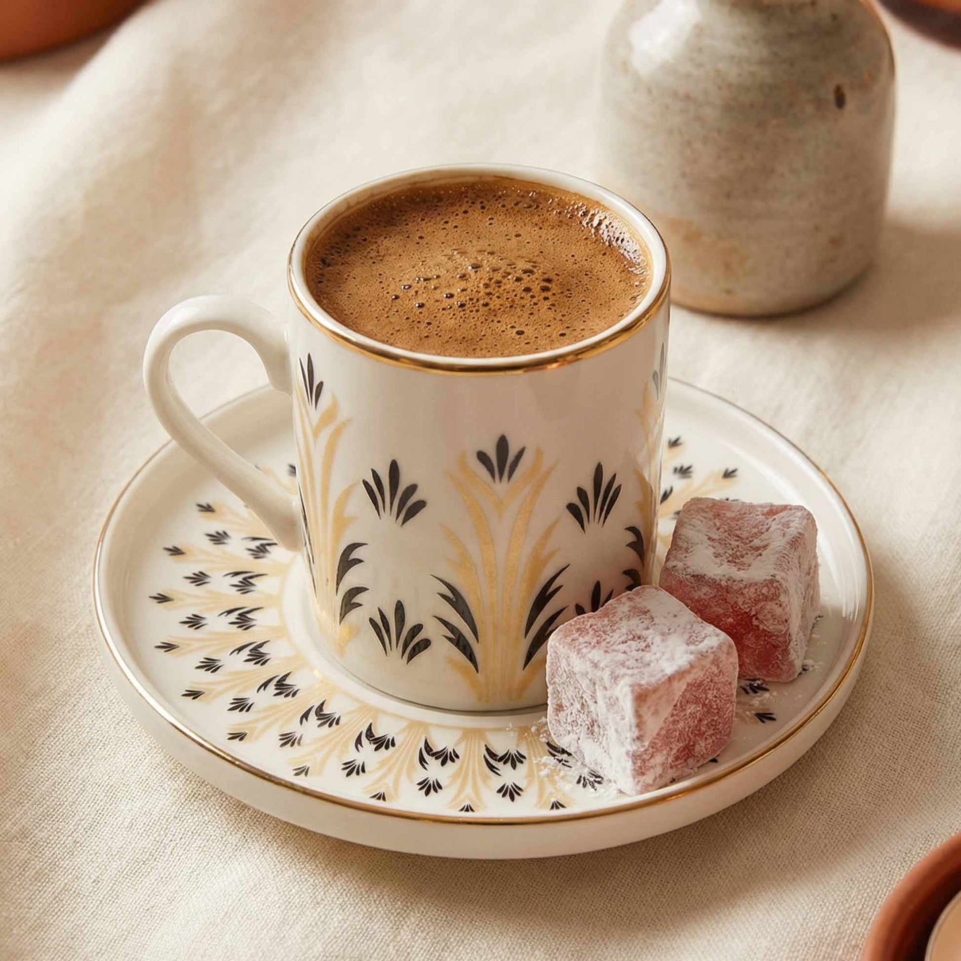 Cup of coffee with a decorative pattern on a matching saucer, accompanied by two pieces of Turkish delight on a beige surface.