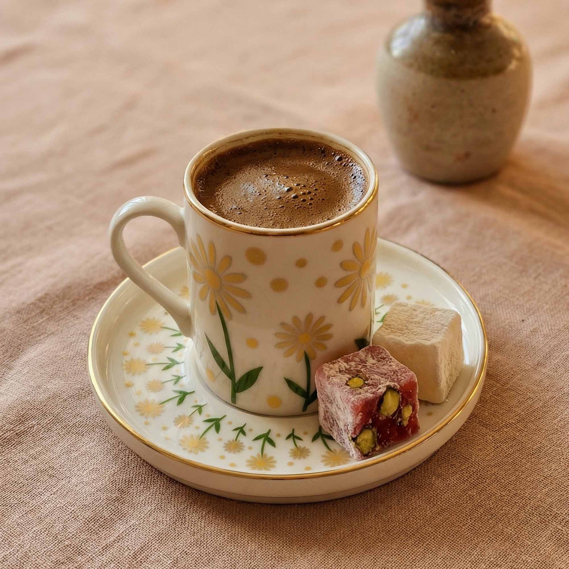 Cup of coffee with a saucer featuring floral designs, accompanied by two pieces of Turkish delight on a textured surface.