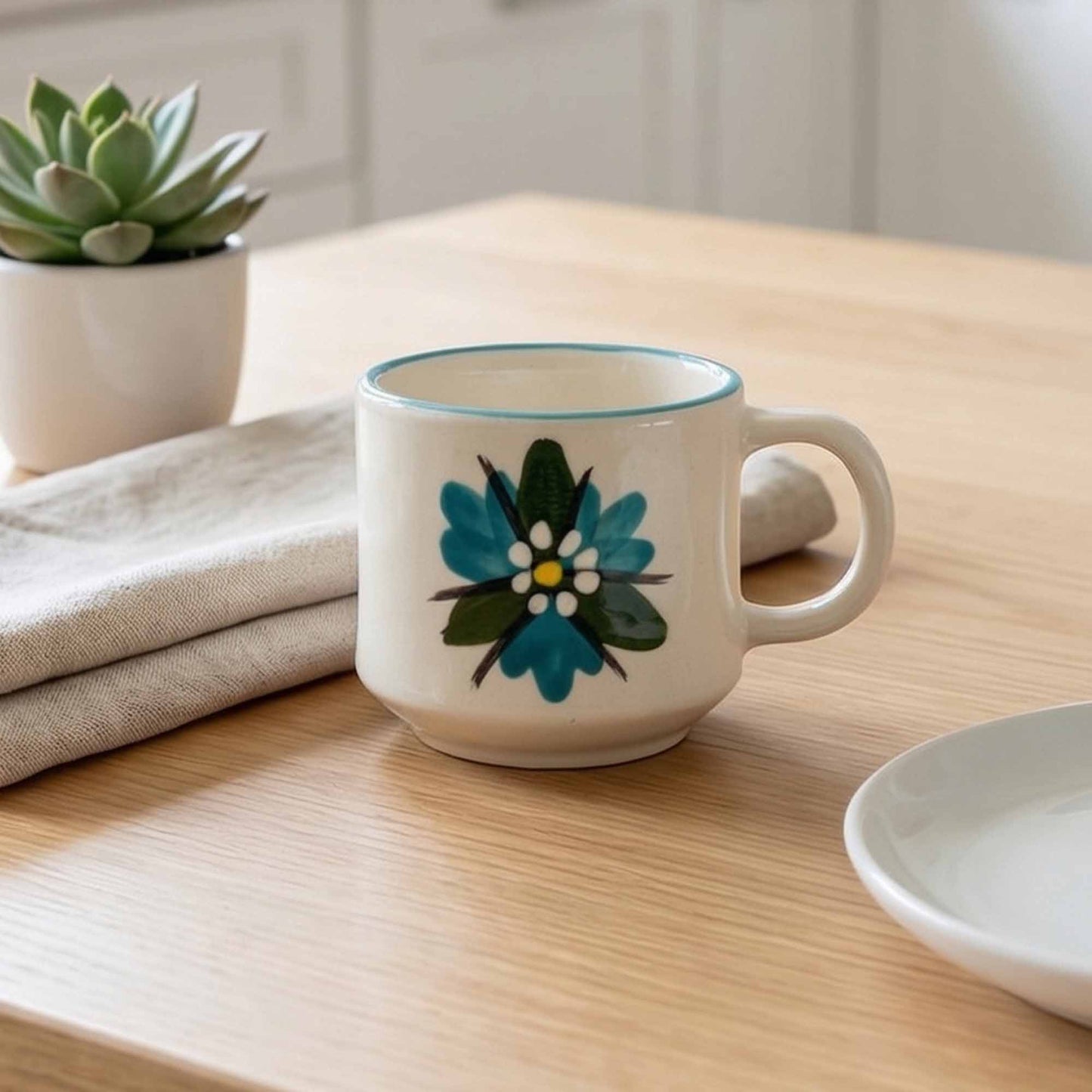 White mug with floral design on a wooden table with a plant and folded cloth in the background
