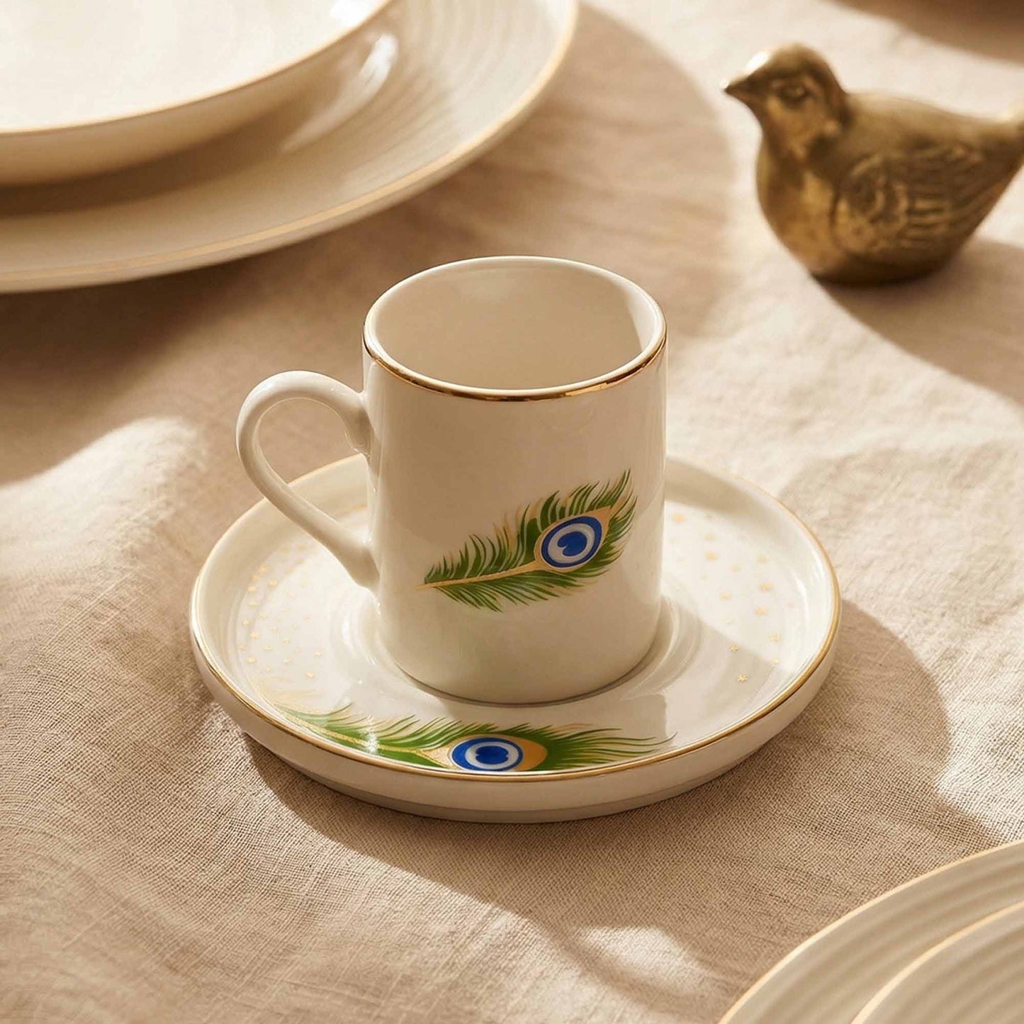 Porcelain cup and saucer with peacock feather design on a beige tablecloth.