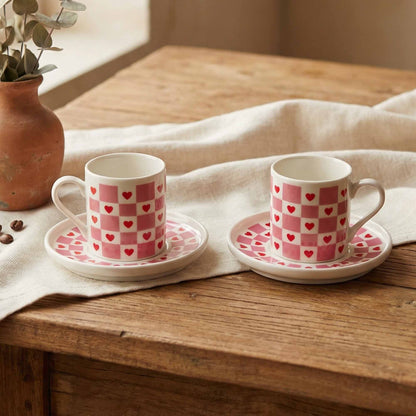 Two porcelain cups with pink checkered and heart patterns on a wooden table with a white cloth.