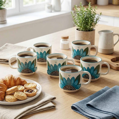 Set of coffee mugs with floral design on a wooden table with pastries and plants.
