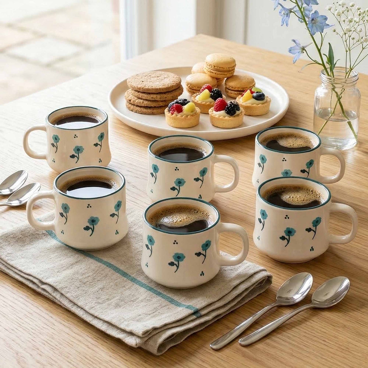 Set of coffee mugs with floral design on a wooden table with pastries and a vase.