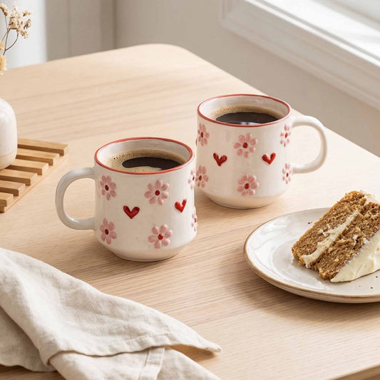 Two mugs with floral designs filled with coffee on a wooden table with a slice of cake.