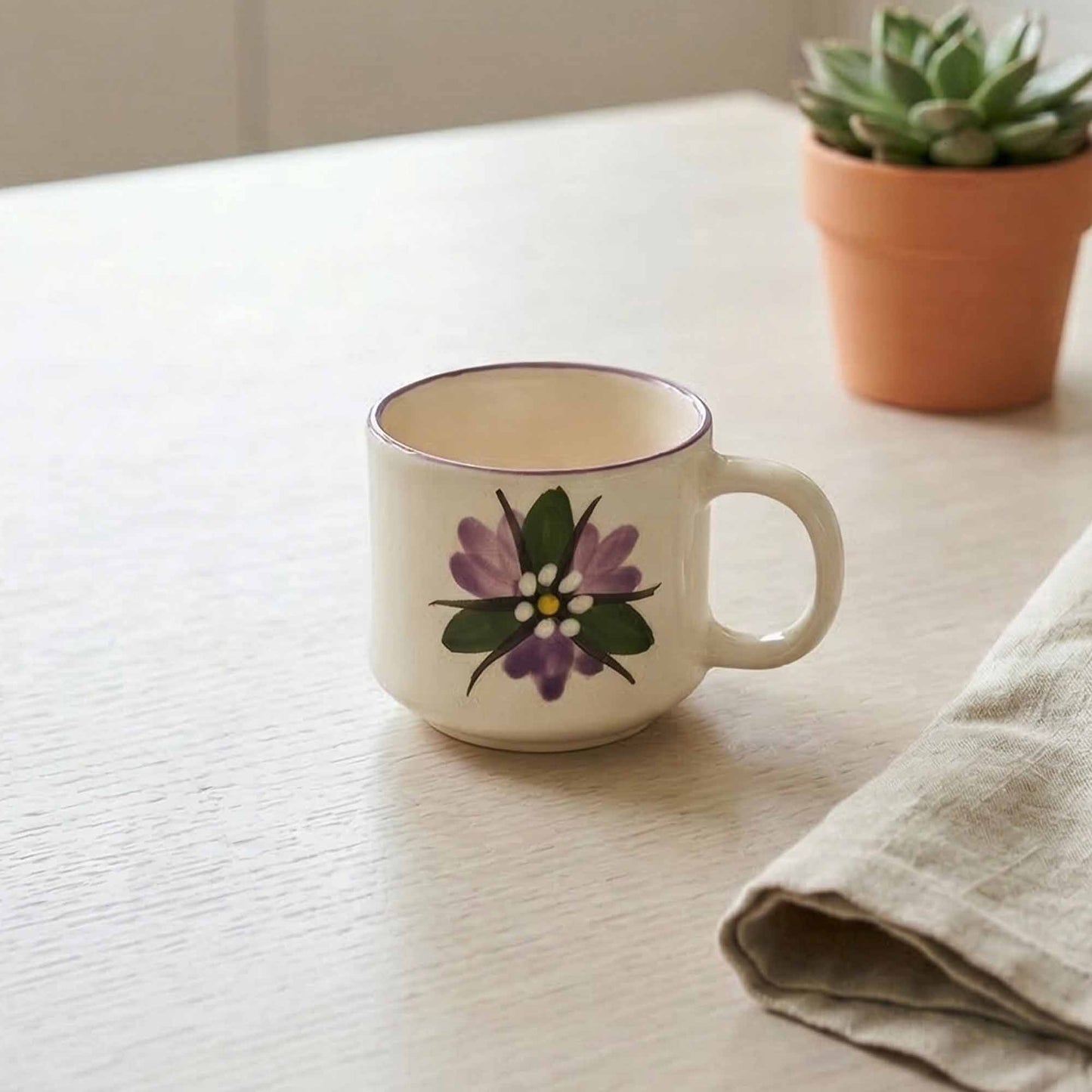 Ceramic mug with floral design on a white surface, next to a potted plant and folded cloth.