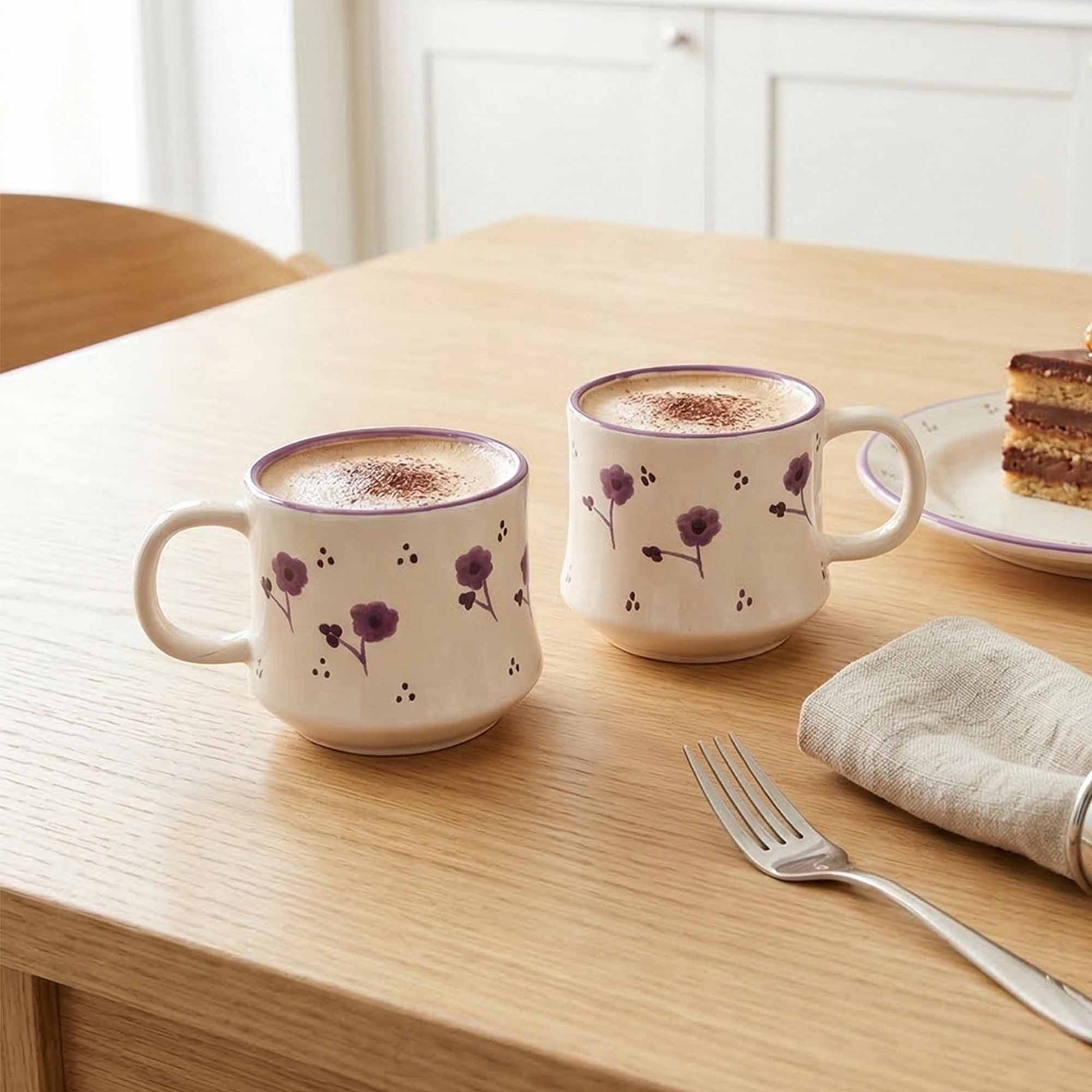 Two floral-patterned mugs with a hot beverage on a wooden table with a plate of cake and fork.