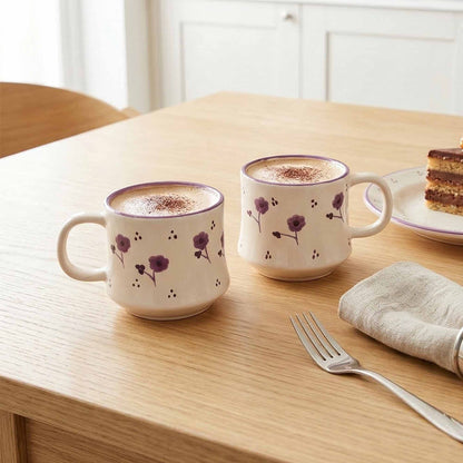 Two floral-patterned mugs with a hot beverage on a wooden table with a plate of cake and fork.