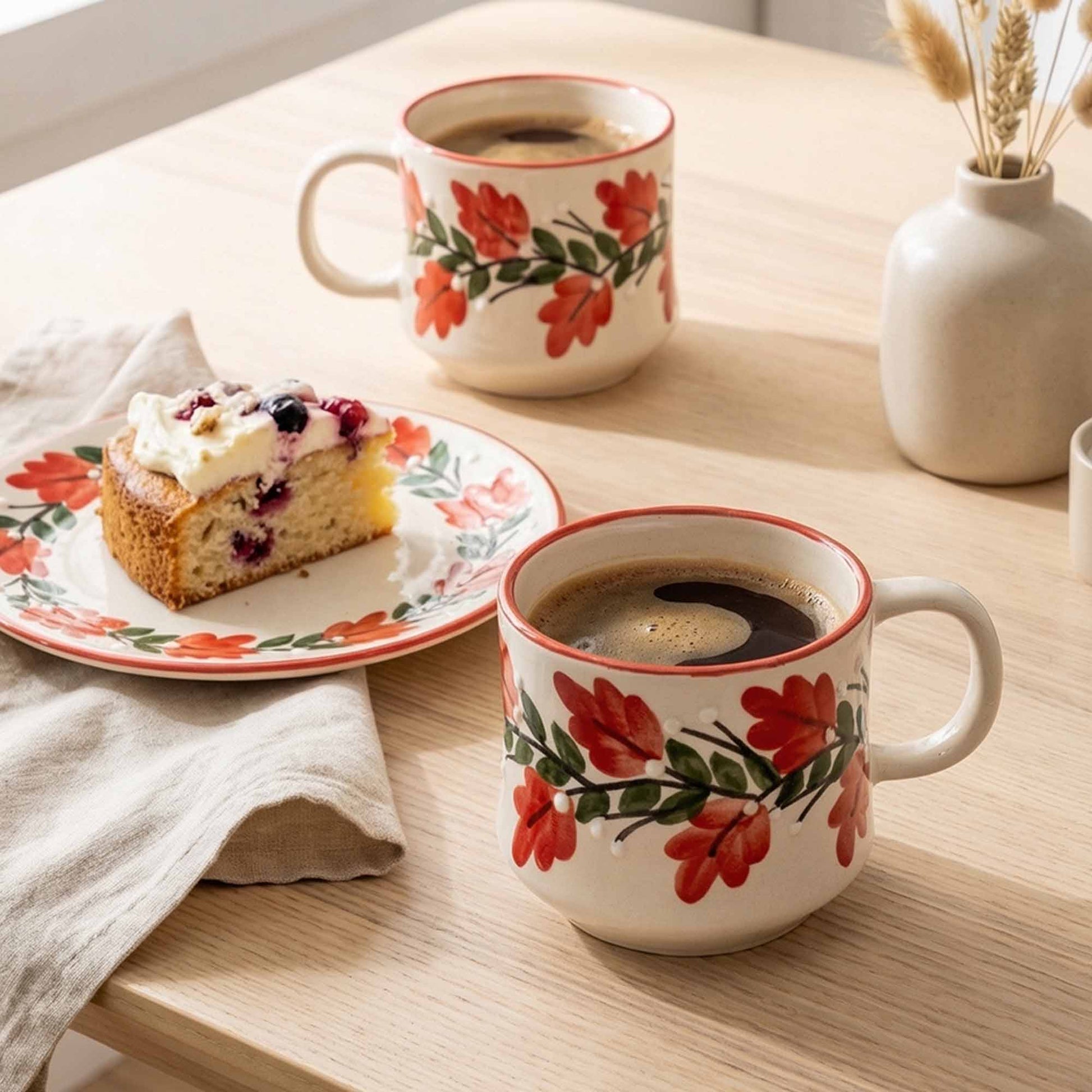Two floral-patterned mugs with coffee and a slice of cake on a wooden table.