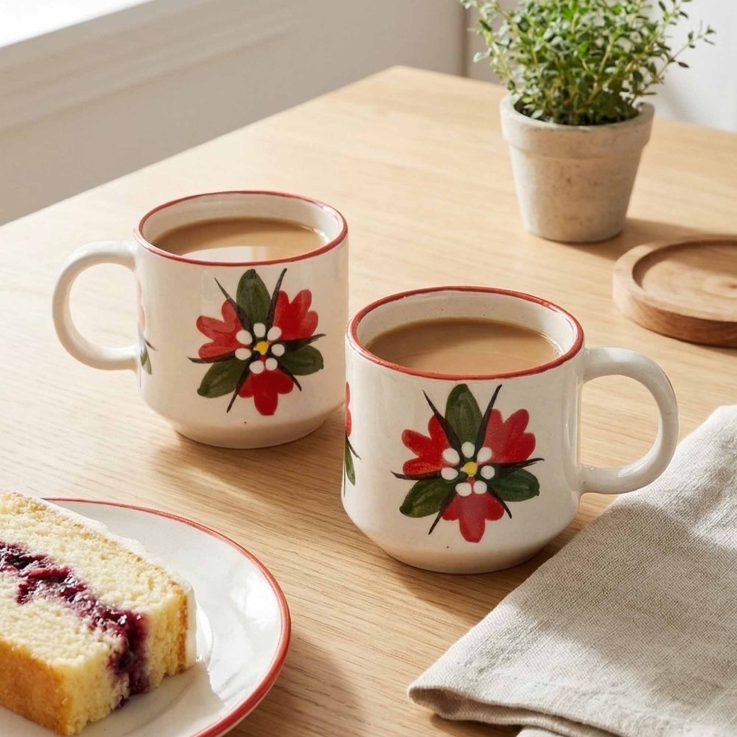 Two floral mugs with coffee on a wooden table with a slice of cake and plant in the background.