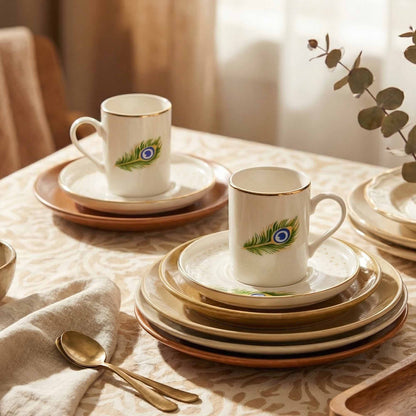 Porcelain cups and saucers with peacock feather design on a table with a cloth and spoons.