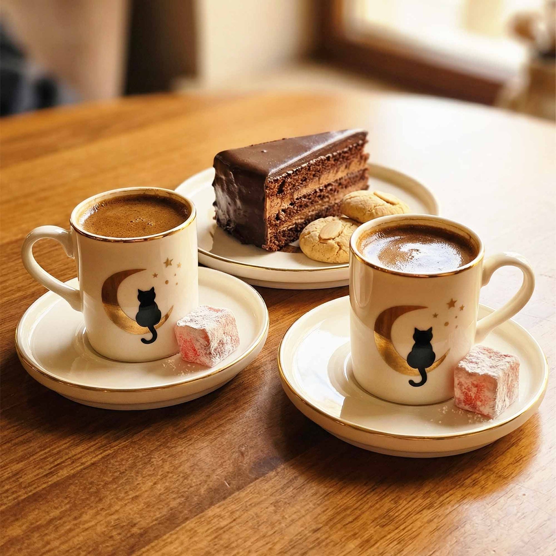 Two cups of coffee with a cat design on saucers, accompanied by a slice of chocolate cake and cookies on a wooden table.