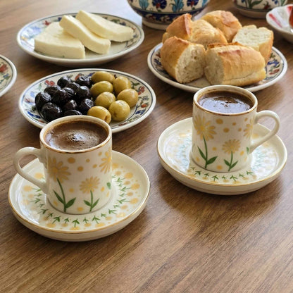 Two cups of coffee with floral designs on saucers, surrounded by bread and olives on a wooden table.