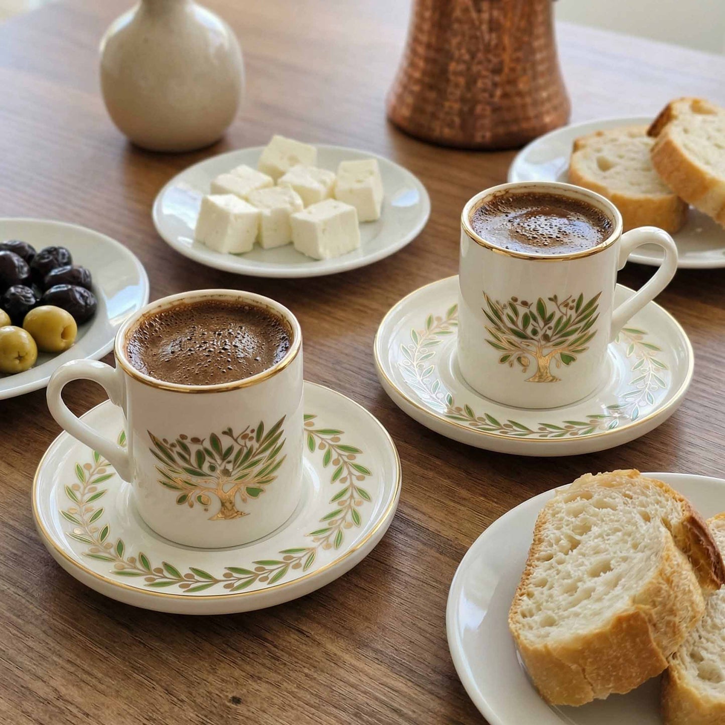 Two cups of coffee with decorative patterns on saucers, surrounded by bread, olives, and dates on a wooden table.