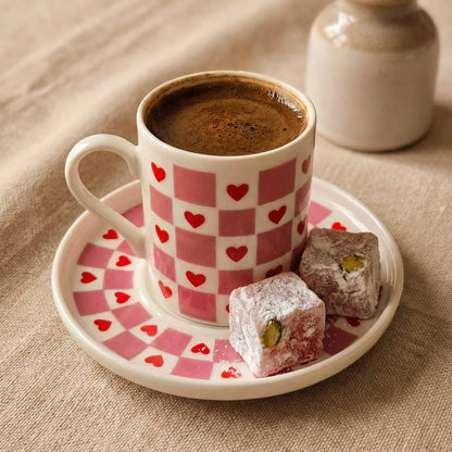 Cup of coffee with a checkered pink and white design on a saucer with heart patterns, accompanied by two pastries on a beige surface.
