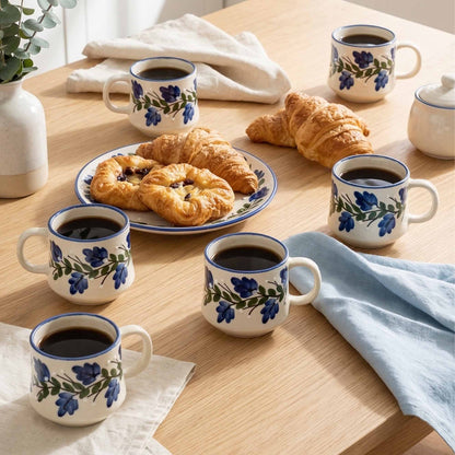 Table setting with floral mugs, pastries, and a plate of bread on a wooden surface.