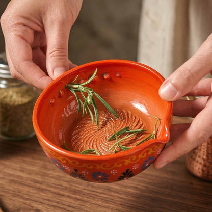 Hand holding an orange ceramic herb stripping bowl with decorative patterns and rosemary leaves on a wooden surface.