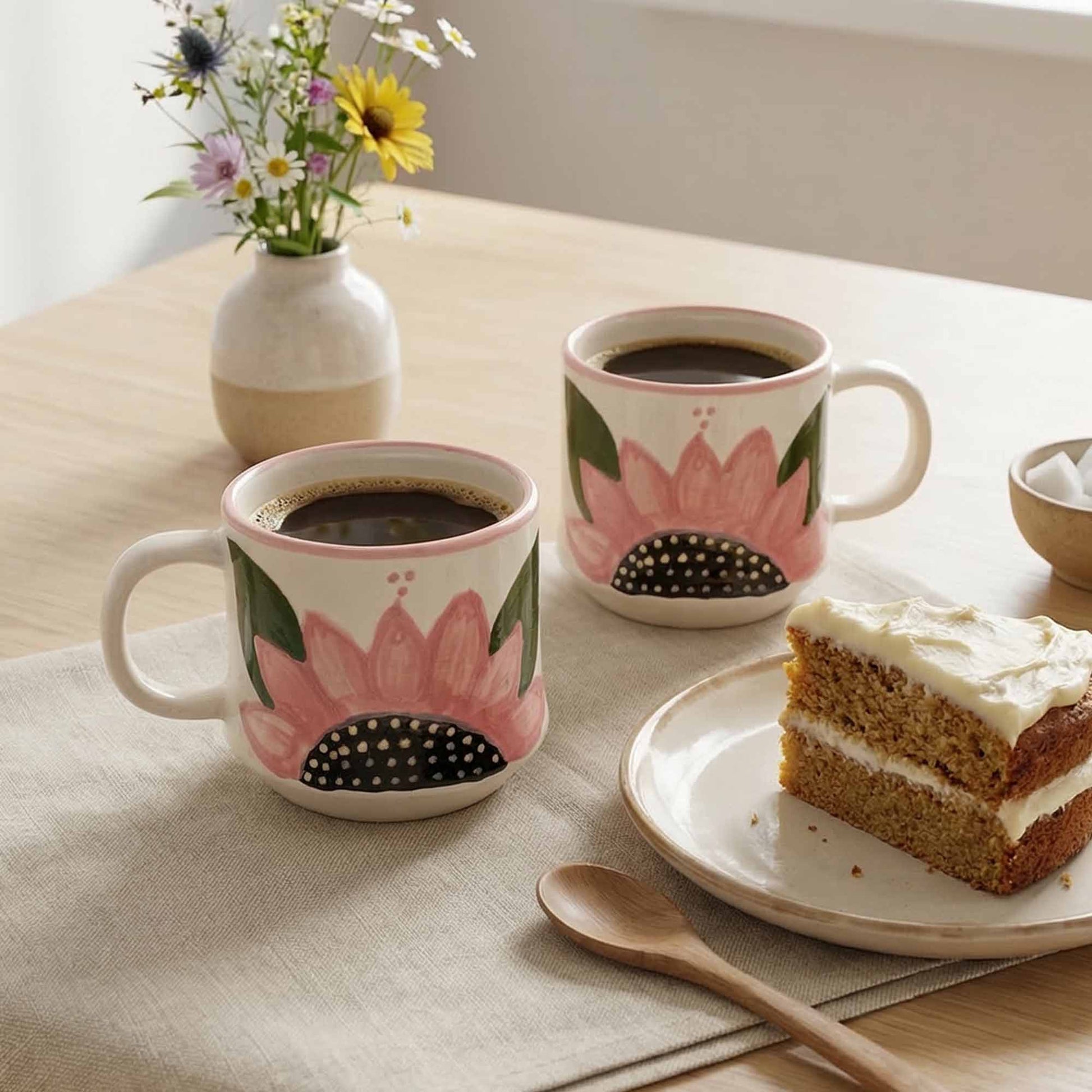 Two floral-patterned mugs with coffee, a slice of cake, and a vase of flowers on a table.
