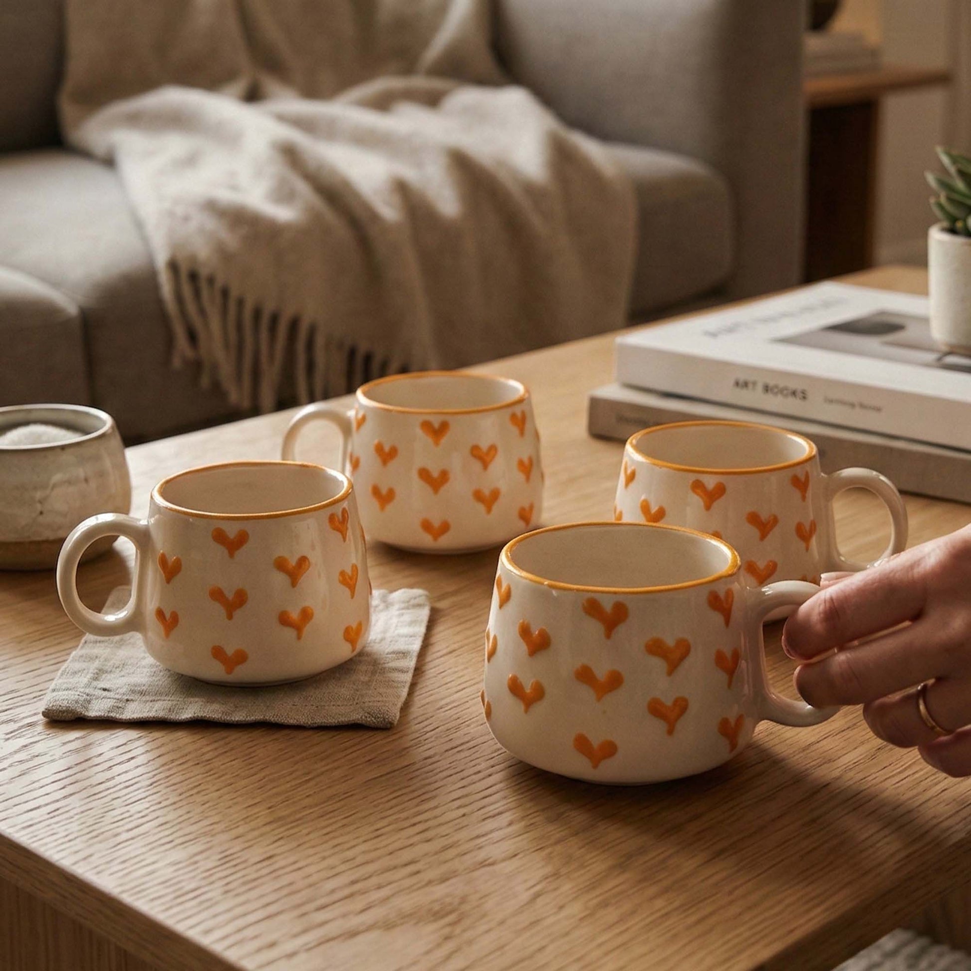 Four ceramic mugs with heart patterns on a wooden table, with a hand picking up one of them.