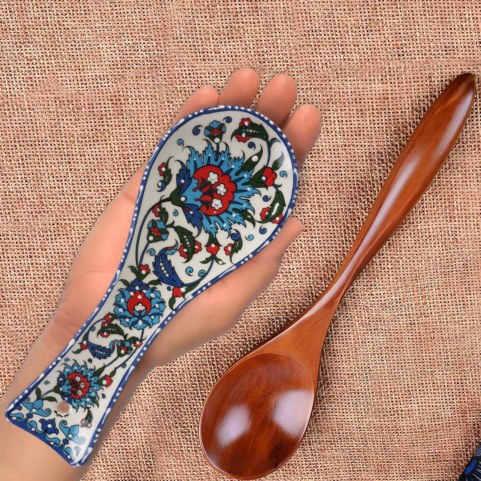 Decorative wooden spoon with a hand holding a ceramic spoon rest featuring intricate floral patterns on a textured fabric background.