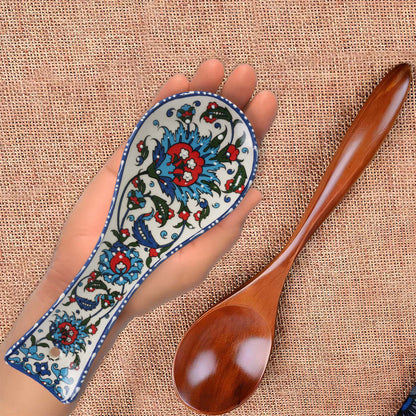 Decorative wooden spoon with a hand holding a ceramic spoon rest featuring intricate floral patterns on a textured fabric background.