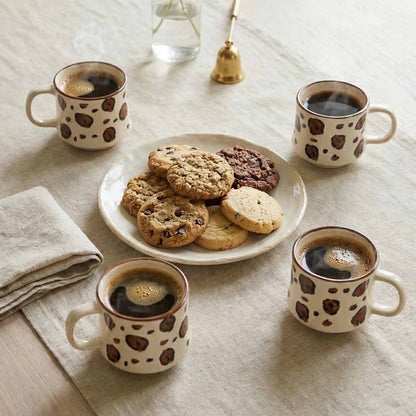 Four mugs with coffee seeds print pattern filled with coffee, around a plate of cookies on a light wooden surface.