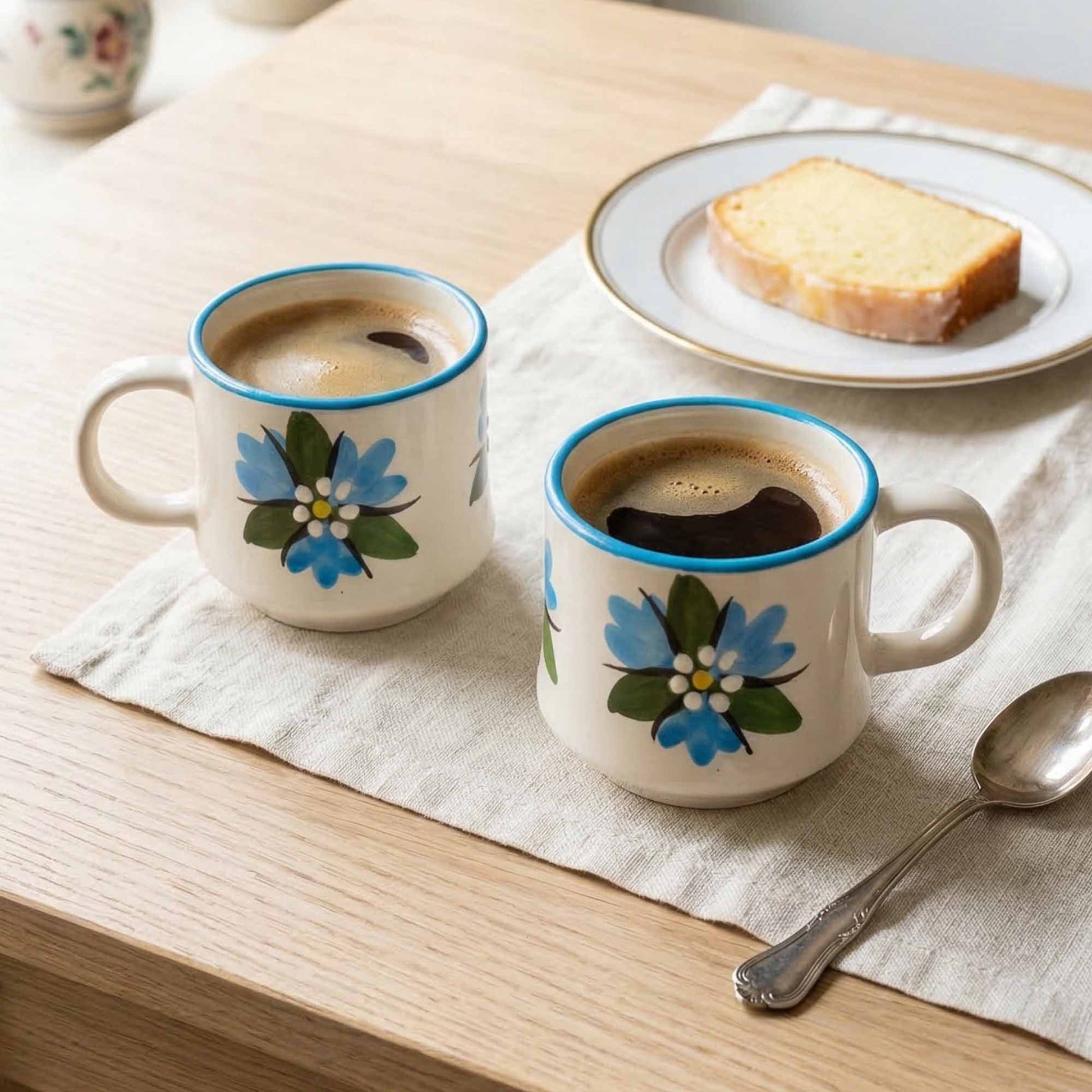 Two floral-patterned mugs with coffee on a wooden table with toast and a spoon.