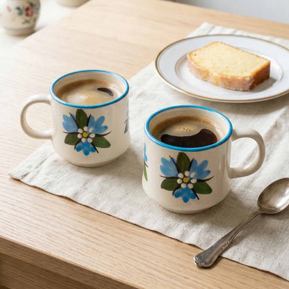 Two floral-patterned mugs with coffee on a wooden table with toast and a spoon.