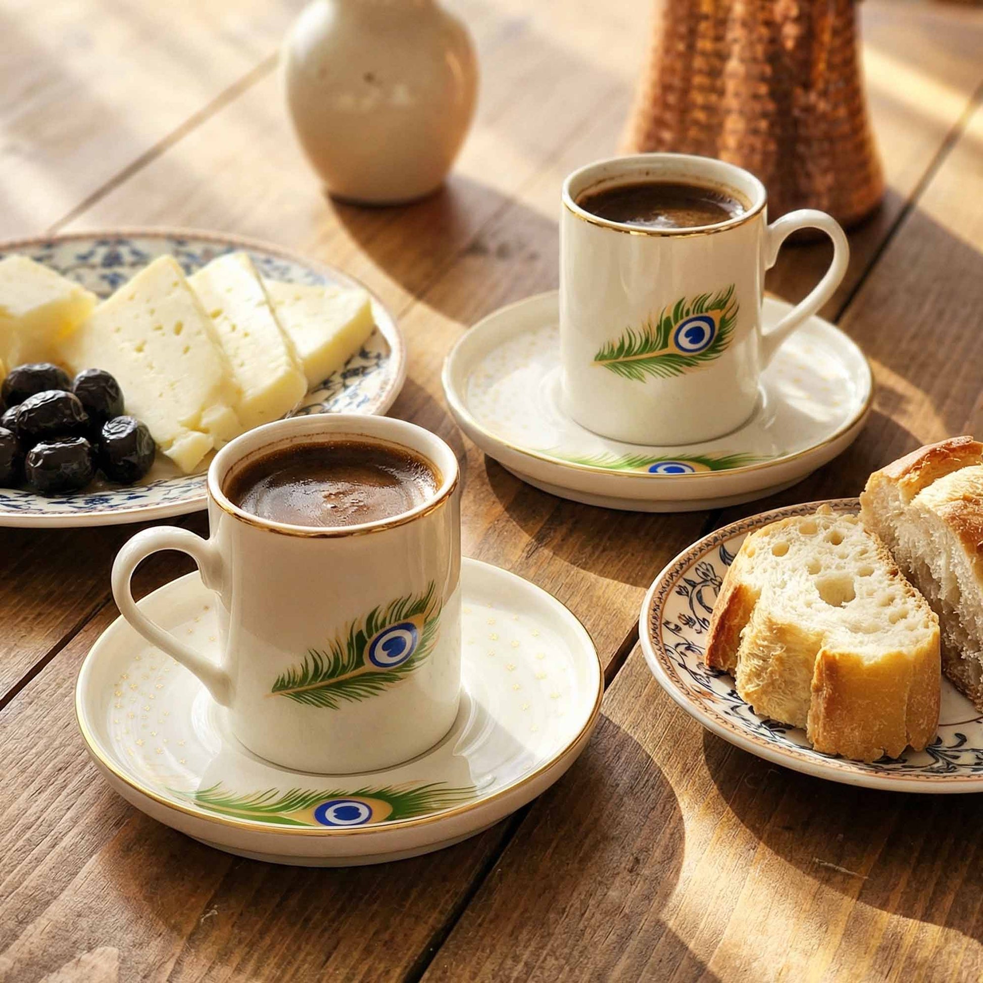 Two cups of coffee with decorative peacock feather designs on saucers, accompanied by bread and cheese on a wooden table.