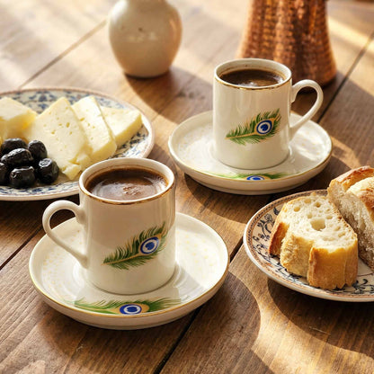 Two cups of coffee with decorative peacock feather designs on saucers, accompanied by bread and cheese on a wooden table.