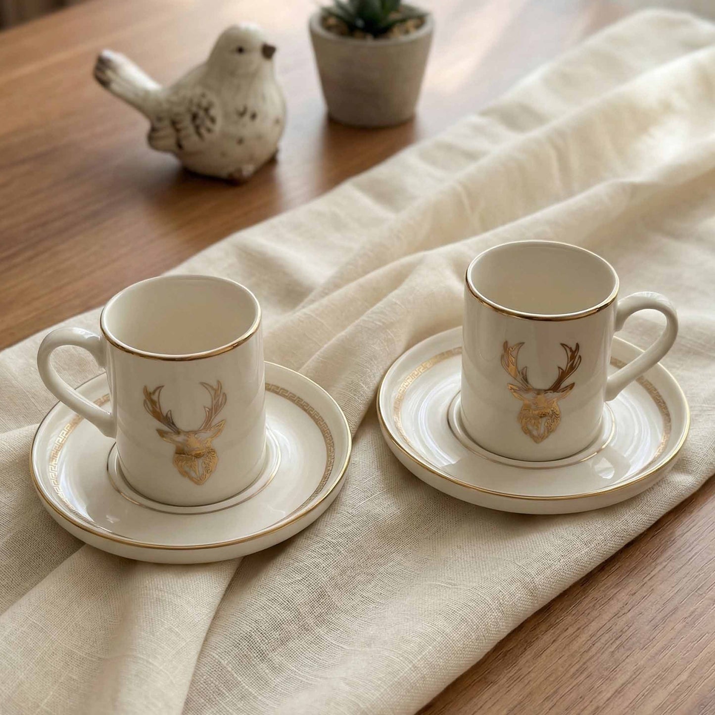 Two porcelain cups with deer head designs on saucers, placed on a wooden table with a white cloth.