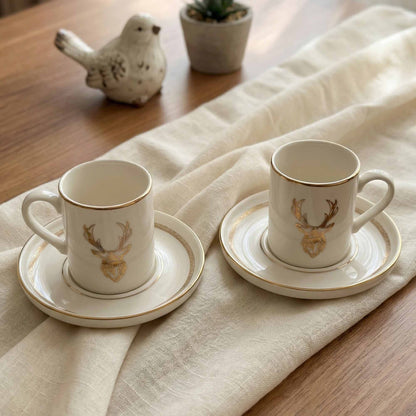 Two porcelain cups with deer head designs on saucers, placed on a wooden table with a white cloth.