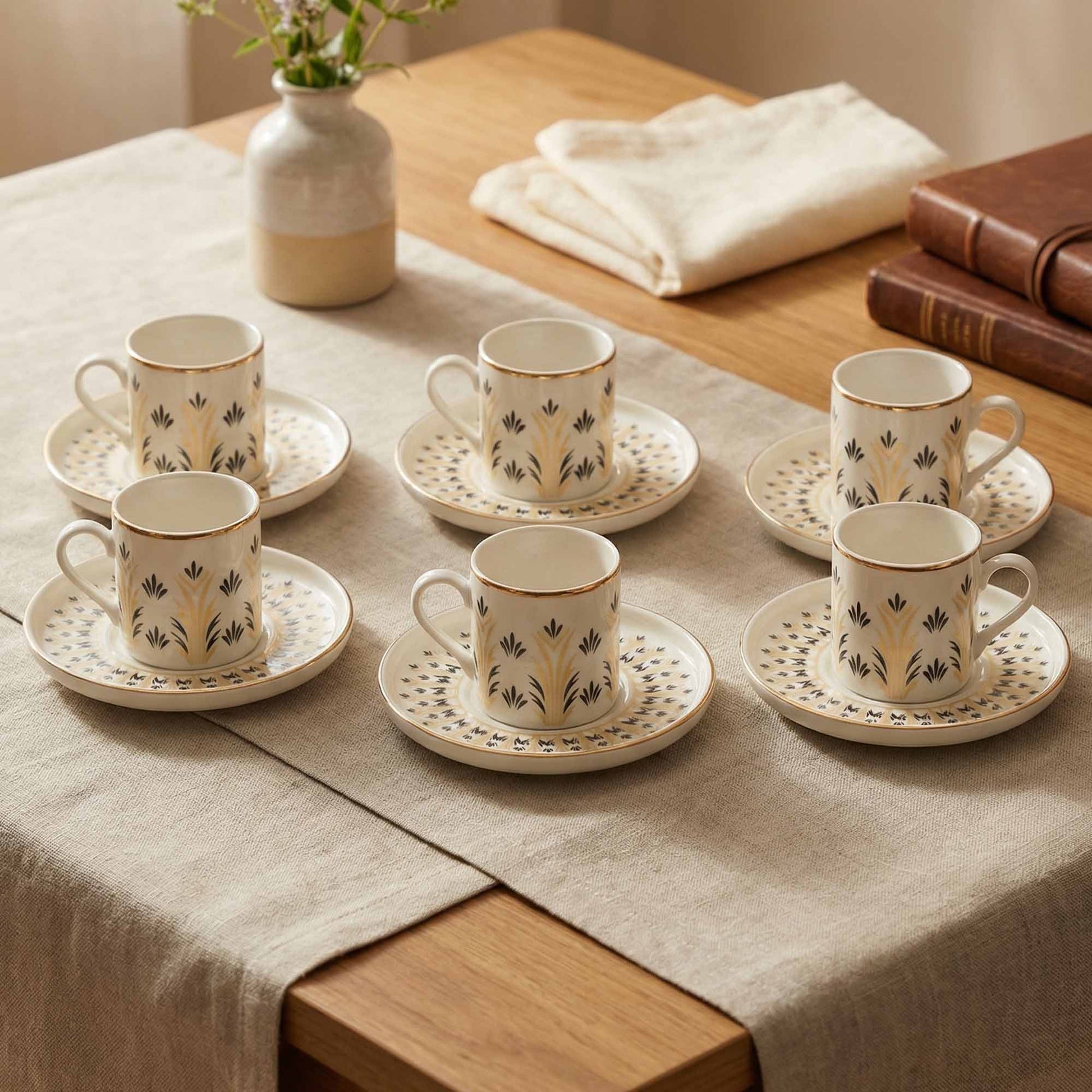 Set of six porcelain cups and saucers on a wooden table with a vase and books in the background.