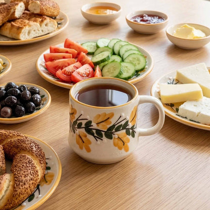 Cup of coffee on a wooden table with plates of bread, fruit, and cheese.