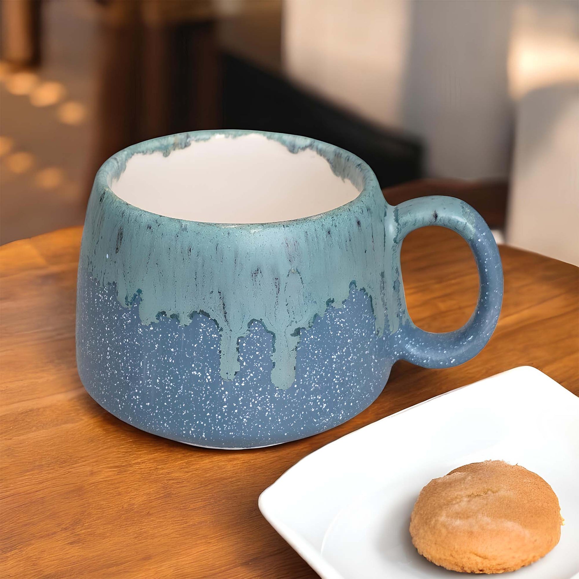 Blue ceramic mug on a wooden table with a cookie on a white plate.