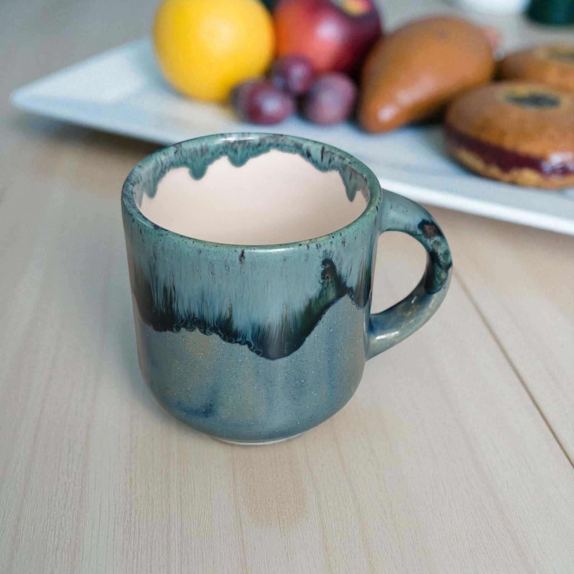 Blue ceramic mug on a wooden surface with a plate of fruit and pastries in the background