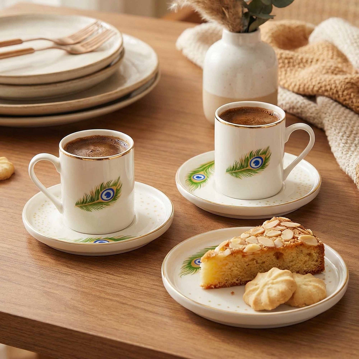 Two cups of coffee with decorative peacock feather patterns on saucers and plates, accompanied by pastries on a wooden table.