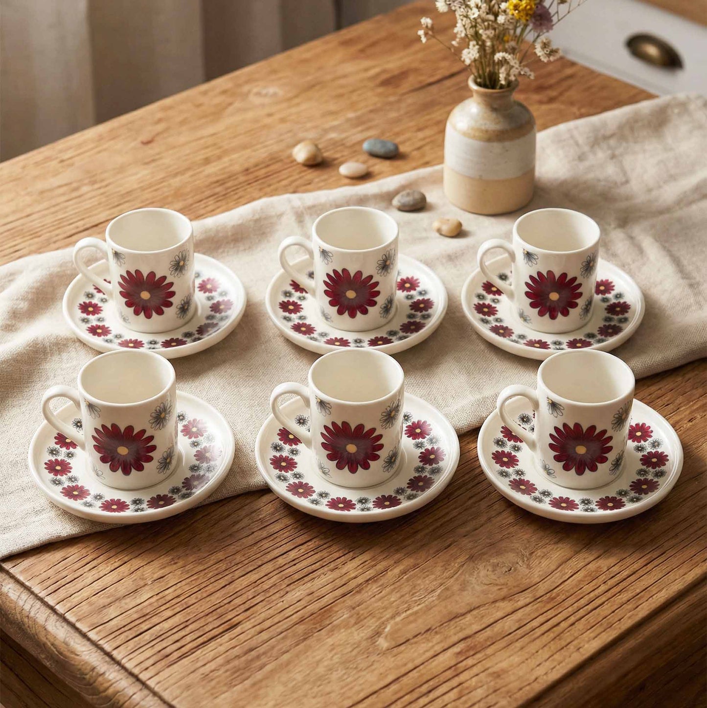 Set of six floral coffee cups and saucers on a wooden table with a vase of flowers in the background.