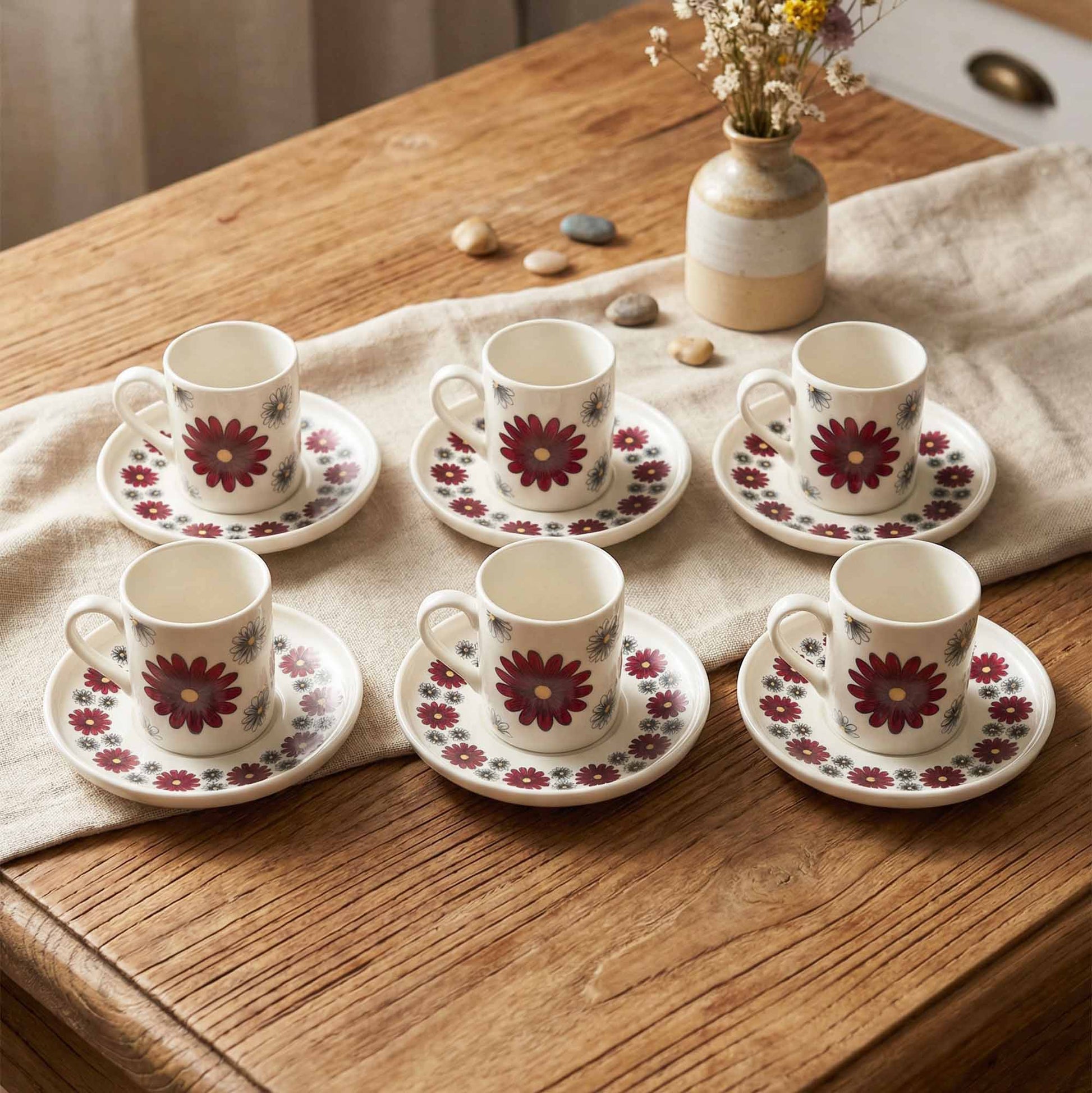 Set of six floral coffee cups and saucers on a wooden table with a vase of flowers in the background.