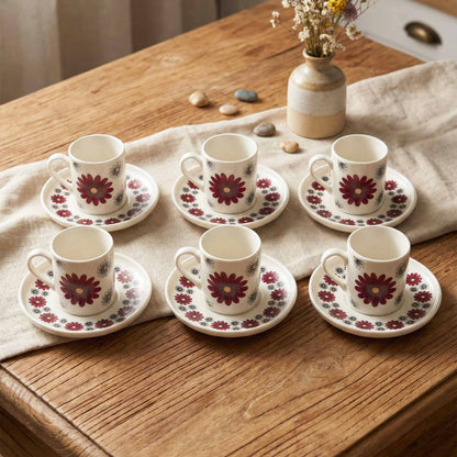 Set of six floral coffee cups and saucers on a wooden table with a vase of flowers in the background.