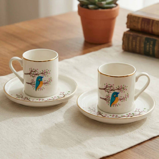 Two porcelain cups with saucers featuring bird designs on a tablecloth.