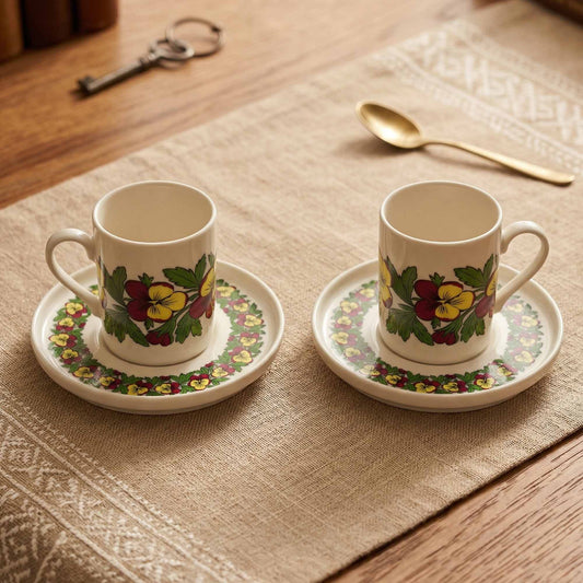 Two porcelain mugs with floral designs on saucers on a textured tablecloth.