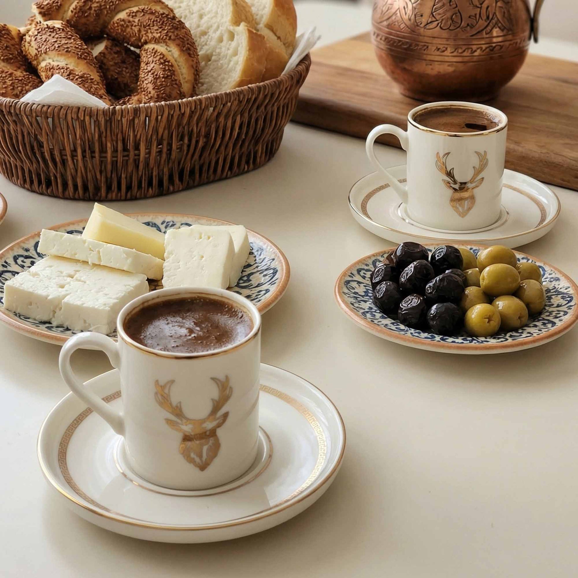 Two cups of coffee with a deer design on saucers, accompanied by bread, cheese, olives, and dates on a table.